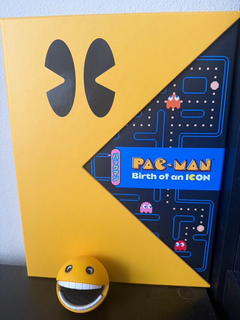 The image features a yellow Pac-Man box with a large black Pac-Man eye design on the upper left corner. A part of the box is cut away in a triangle shape, revealing a black background with a blue maze design, reminiscent of the Pac-Man game, along with the phrase 