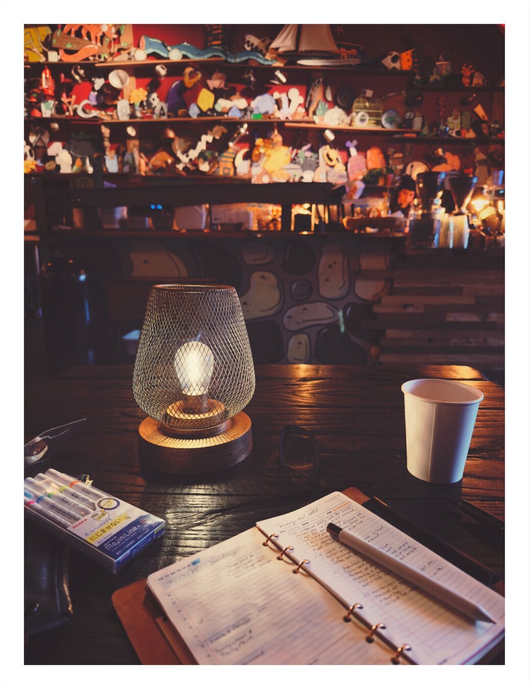 A cozy workspace with an open notebook, pen, markers, and a disposable coffee cup on a wooden table. In the background, shelves are lined with various figurines and memorabilia under warm ambient lighting. A decorative table lamp with a wireframe shade.