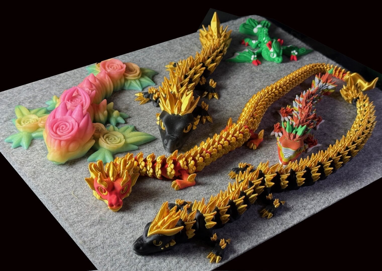 6 3D printed dragons. Upper left: rose printed in multicolor fluorescent filament, next, a black and gold crystal dragon (gold crystals), behind that, a christmas dragon, green, white and red, then a New Year's dragon, gold and red, a poop (multicolored from waste) dragon and finally another, longer, crystal dragon.