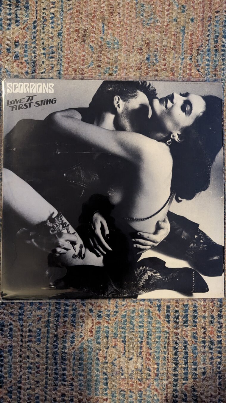 Blank and white vinyl cover of a man and woman hugging and the man has a tattoo needle on her leg. Title of Love At First Sting by Scorpions.