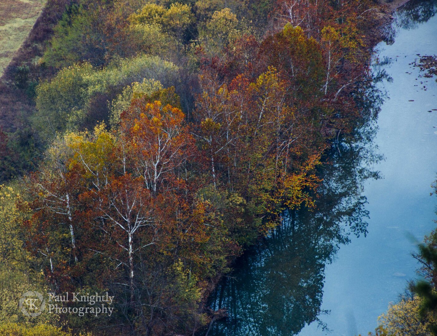 A view looking down onto a river from above. The waters are a vibrant shade of light blue, reflecting the sky and mineral content of the water. The river is bound by trees sporting full fall foliage in colors ranging from red, orange, yellow, and green.