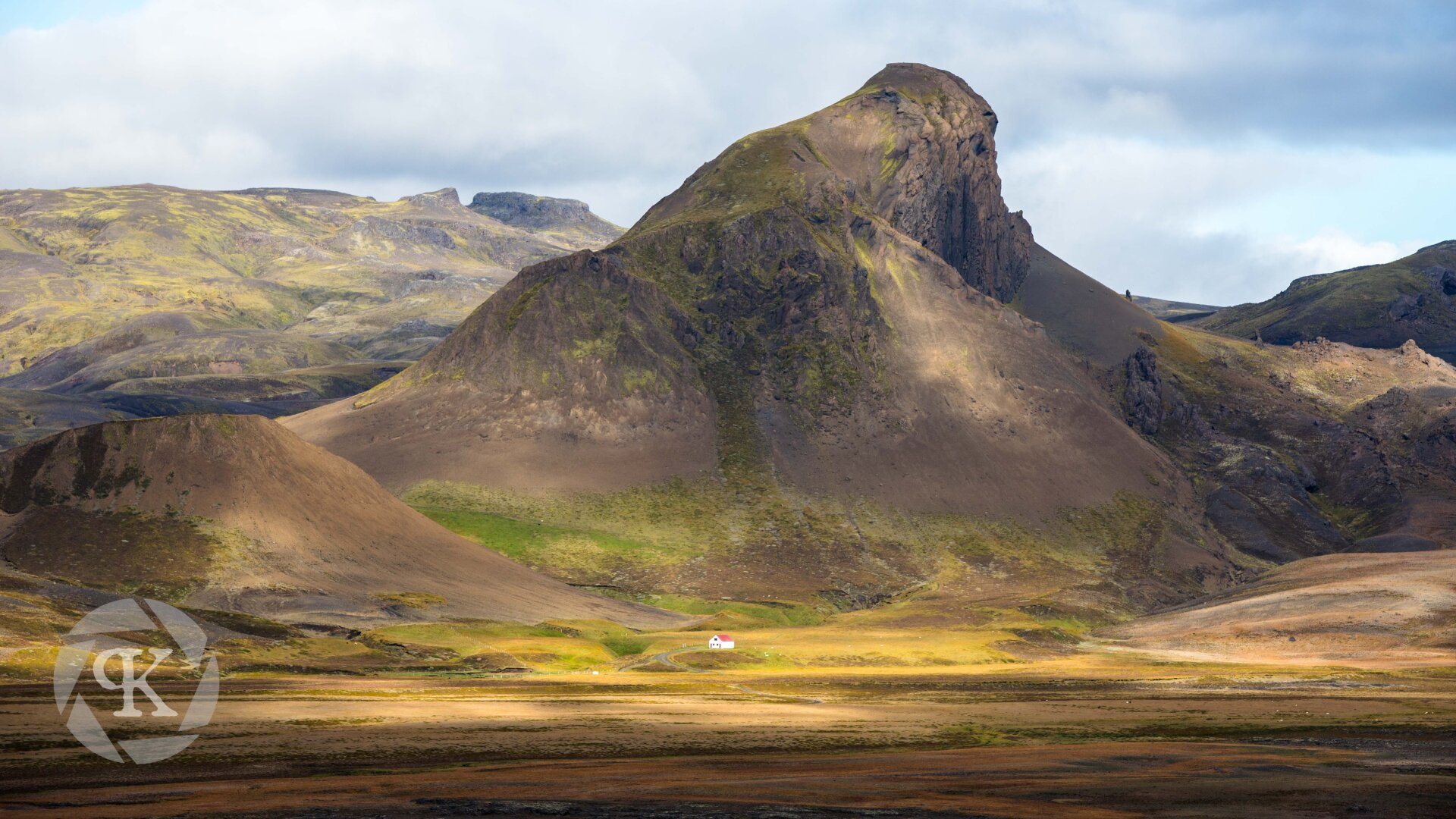 A sweeping view of a golden Icelandic landscape. A large, prominent mountain is situated in center frame with an imposing and almost impossible-seeming steep slop near its summit. A small white house with a red roof is seen below the slope of the mountain, providing a sense of scale for the vastness of the landscape.