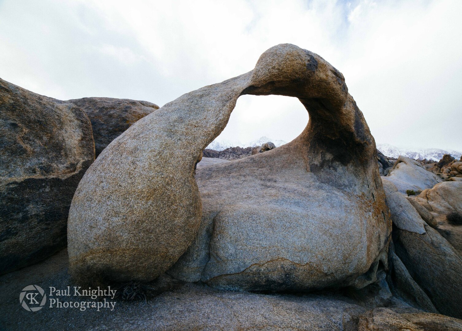 A rock formation that has been eroded out in the center by wind and rain, this arch takes on the form of a Möbius strip with a two prominent lobes near its base. The formation is set against a pale sky and stark shadows among the crevices of the boulders.