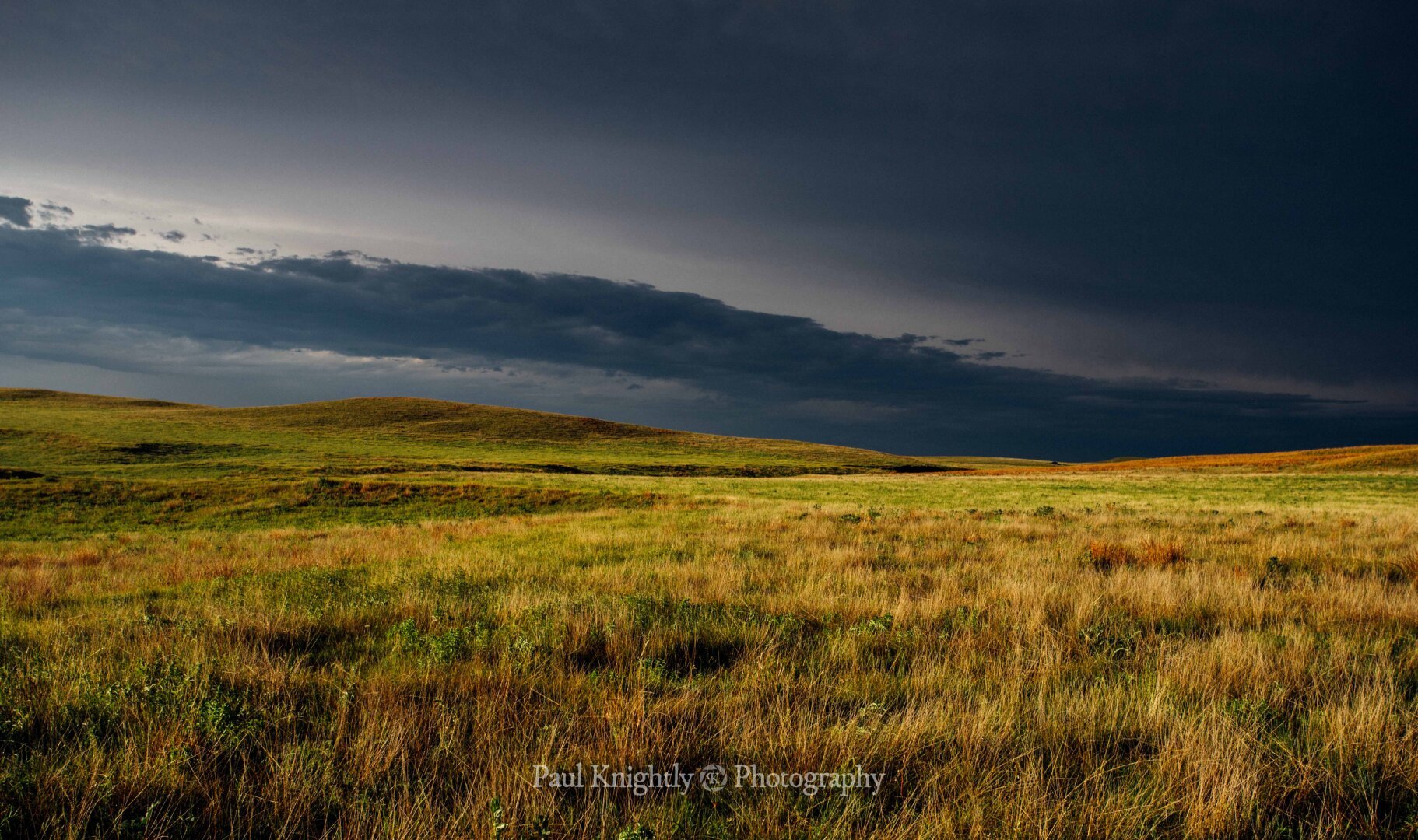 Rolling hills of golden and green grass stretch to the horizon. The illuminated hills sit beneath a dark and ominous sky on the backside of a thunderstorm. The clouds are accentuated by varying shades of dark blue to white.