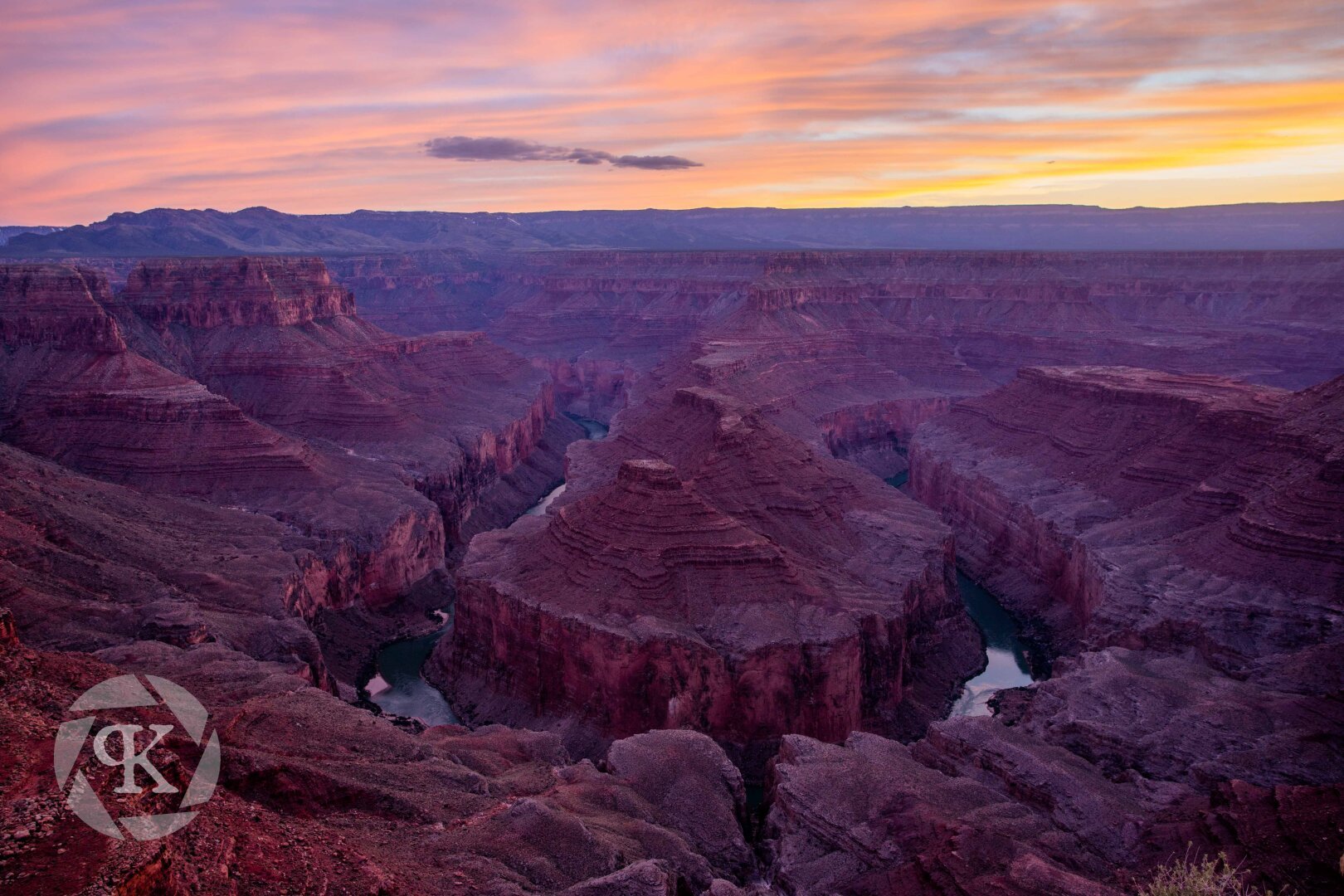 A horseshoe-type bend along a river carving out a deep section of canyon. The landscape is bathed in the red and yellow glow of the setting sun under a vibrant sunset sky illuminated by high altitude clouds.