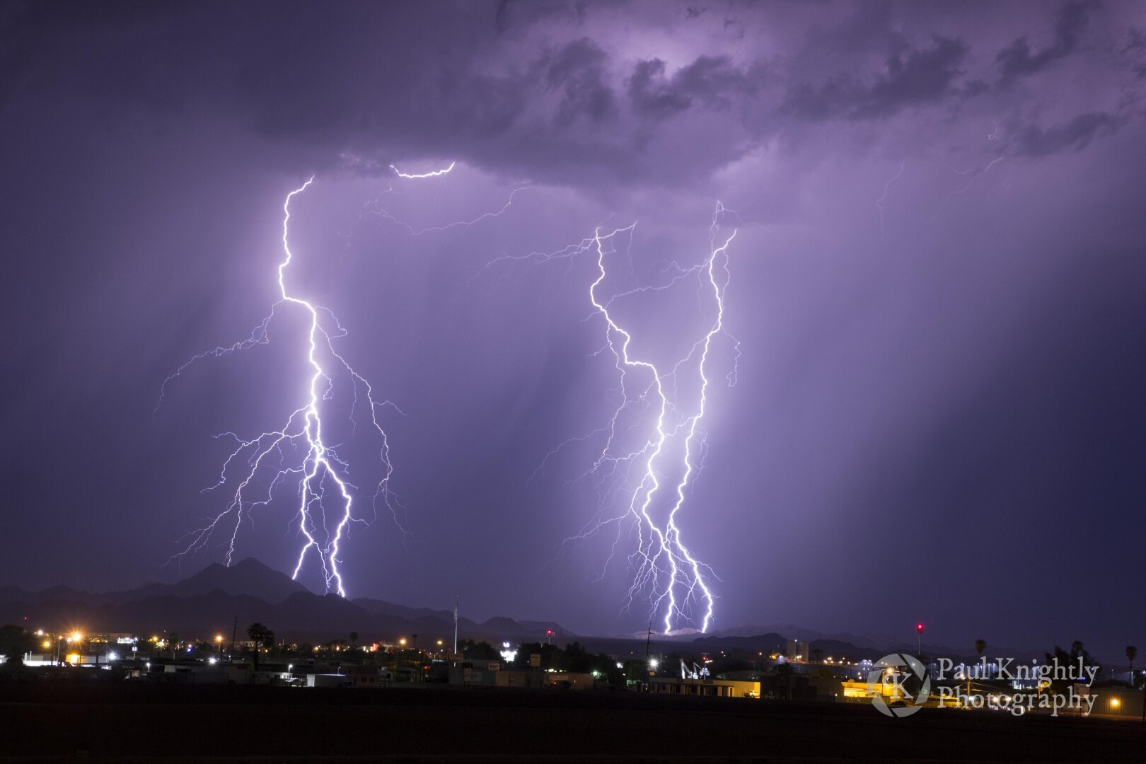 Three cloud to ground bolts of lightning are in center frame of this image, illuminating curtains of rain and showing the full body of the bolts from the base of the cloud to where they land in distant mountains. The lights of Yuma, Arizona dot the foreground of the scene.