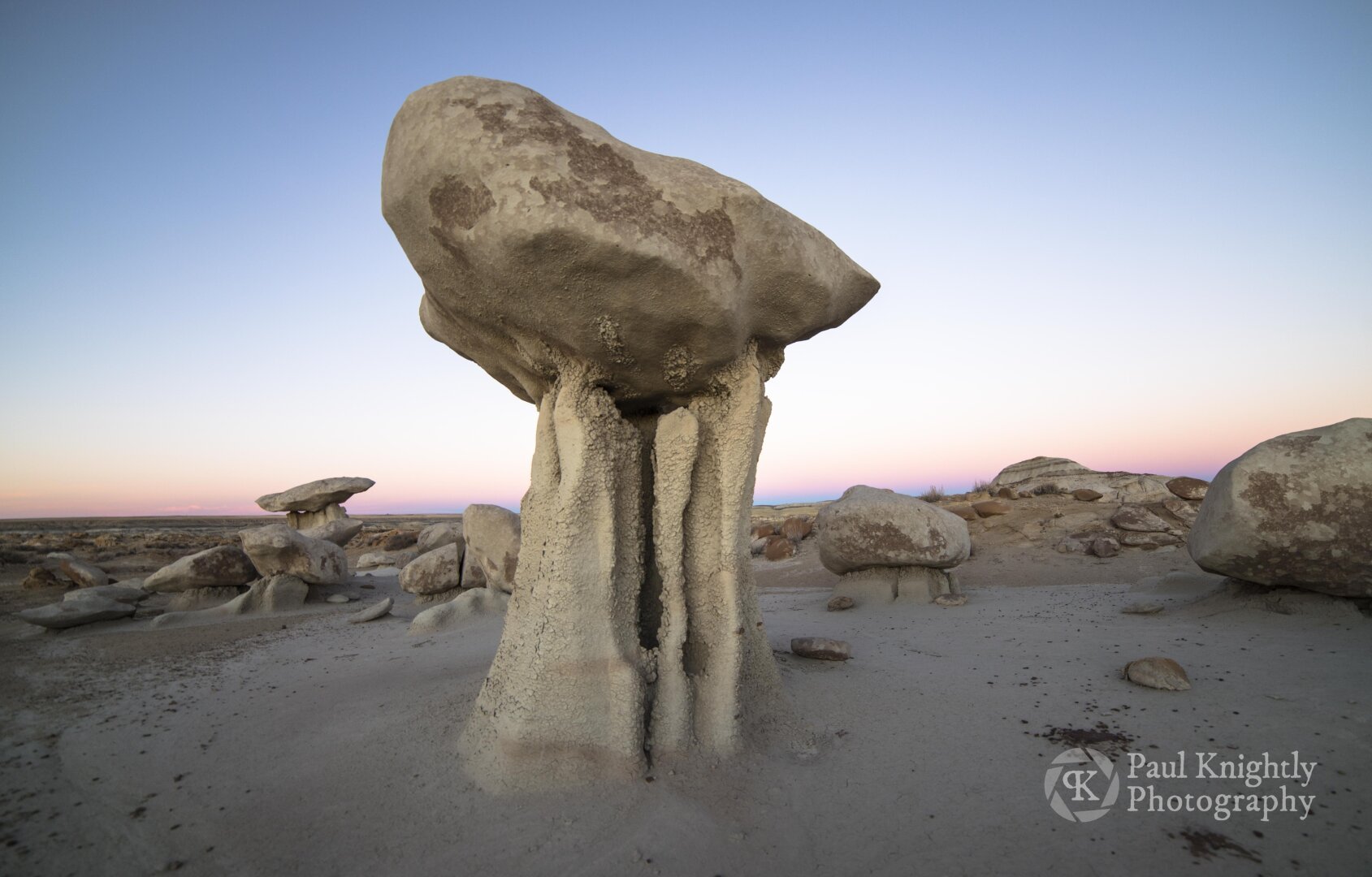 A delicate hoodoo stands guard over a desert landscape. The hoodoo consists of a large boulder poised on top of a narrow column of mudstone, eroded by wind and rain but preserved by the relative dryness of the region. The blue light afterglow of the sky is defined by the advancing Belt of Venus, a narrow band of pink light, along the eastern horizon.