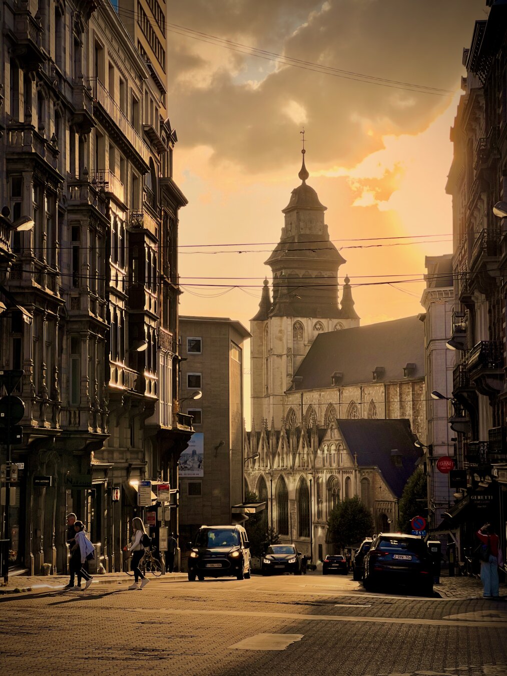 A narrow city street is framed in a tall “canyon” composition, with old stone buildings lining both sides and a church rising in the center distance. Warm golden-hour sunlight breaks through clouds, casting long shadows and highlighting the church’s gray domed roof and slender spire, while overhead power lines cut across the sky. A few pedestrians and parked cars line the street at the bottom of the frame, creating a calm, nostalgic atmosphere.