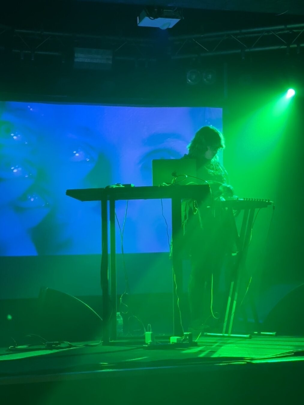 Cholly on stage with electronic gear in front, lit in blue with video projection behind