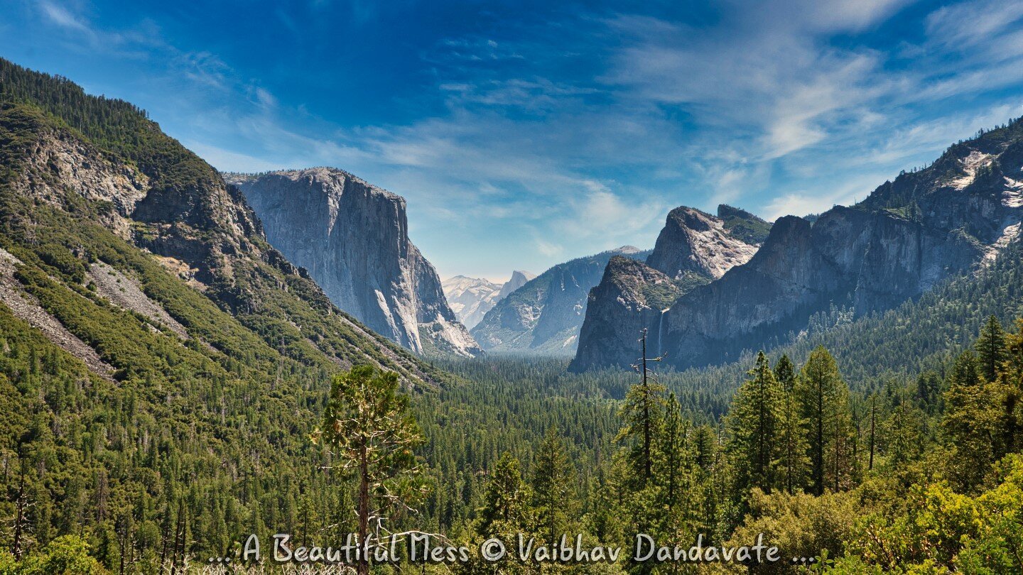 A stunning view of Yosemite Valley with lush green forests, towering granite cliffs, and a clear blue sky. The iconic El Capitan and Half Dome are visible in the distance, showcasing the grandeur of this natural wonder.