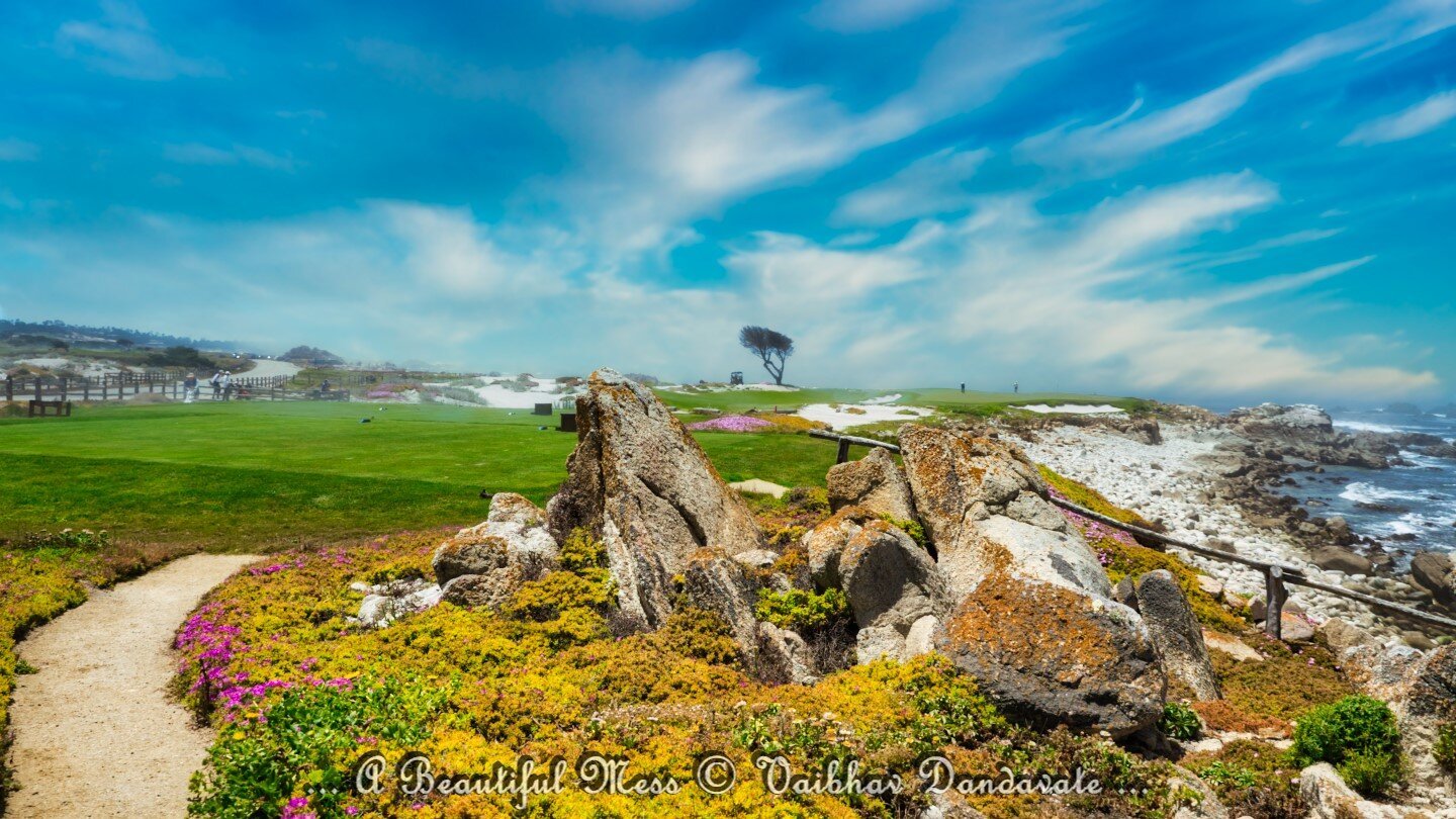 A picturesque view of a golf course on the 17-Mile Drive in Carmel-by-the-Sea, California. The image features a rocky foreground, lush green fairways, and the ocean in the background under a bright blue sky.