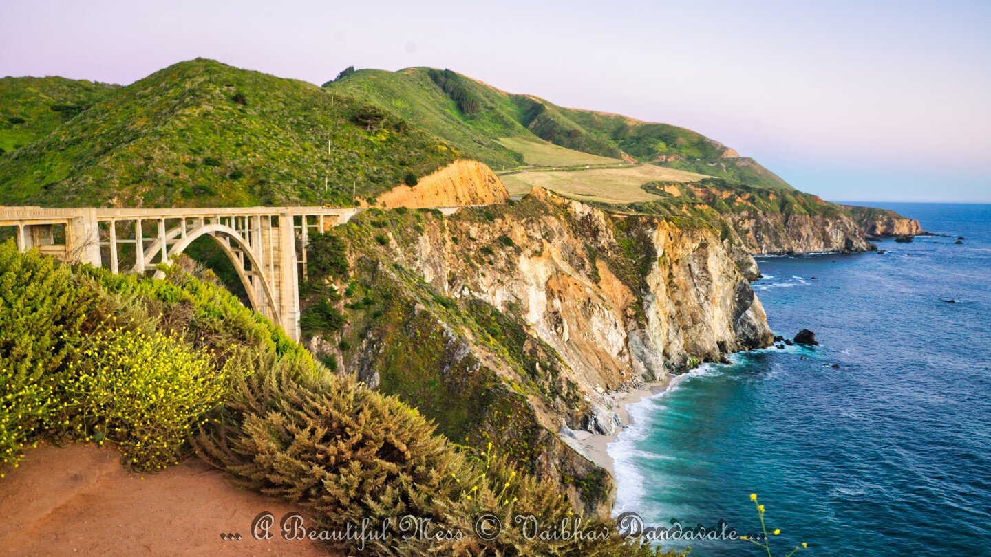 A stunning view of Big Creek Bridge on US Coastal Highway 1 at Big Sur, California. The bridge spans a deep gorge with lush green hills in the background and waves crashing against the cliffs below. Captured by Vaibhav Dandavate.