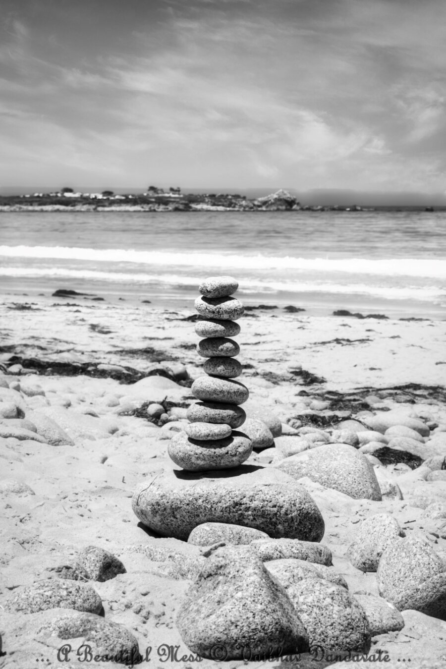 A black-and-white image of a stack of smooth stones balanced on a sandy beach with the ocean and rocky shoreline in the background.