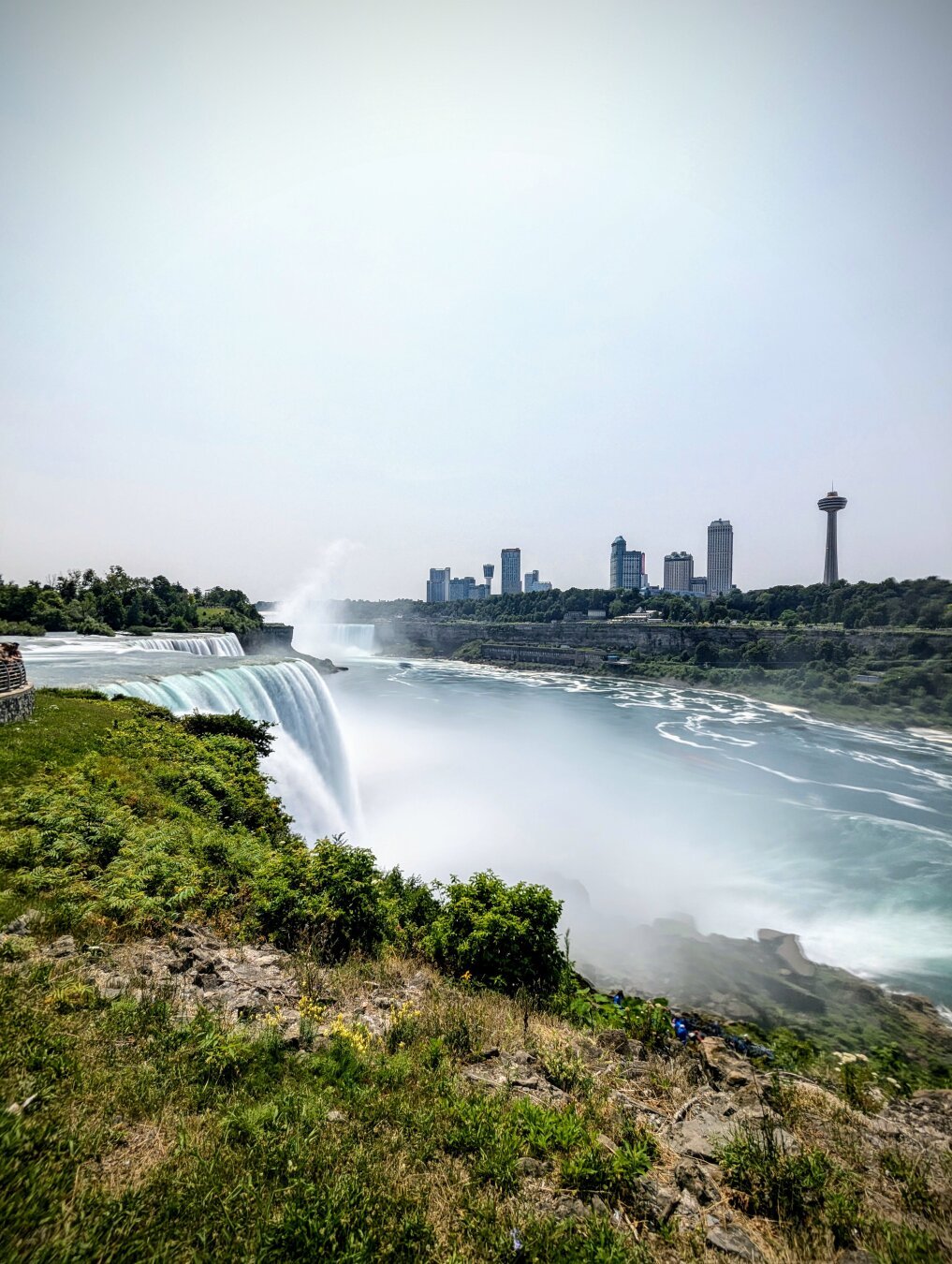 A stunning view of Niagara Falls with water cascading down the cliffs, surrounded by lush greenery and a distant city skyline under a clear sky.