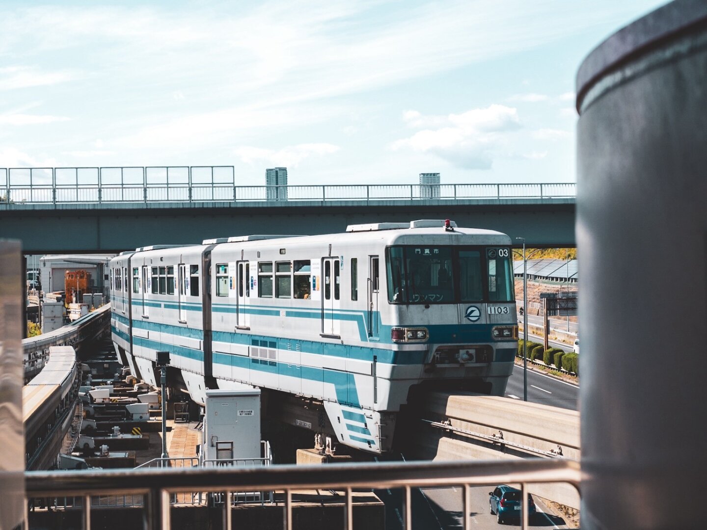 A light blue and white monorail train traveling on elevated tracks with a highway and overpass visible in the background.