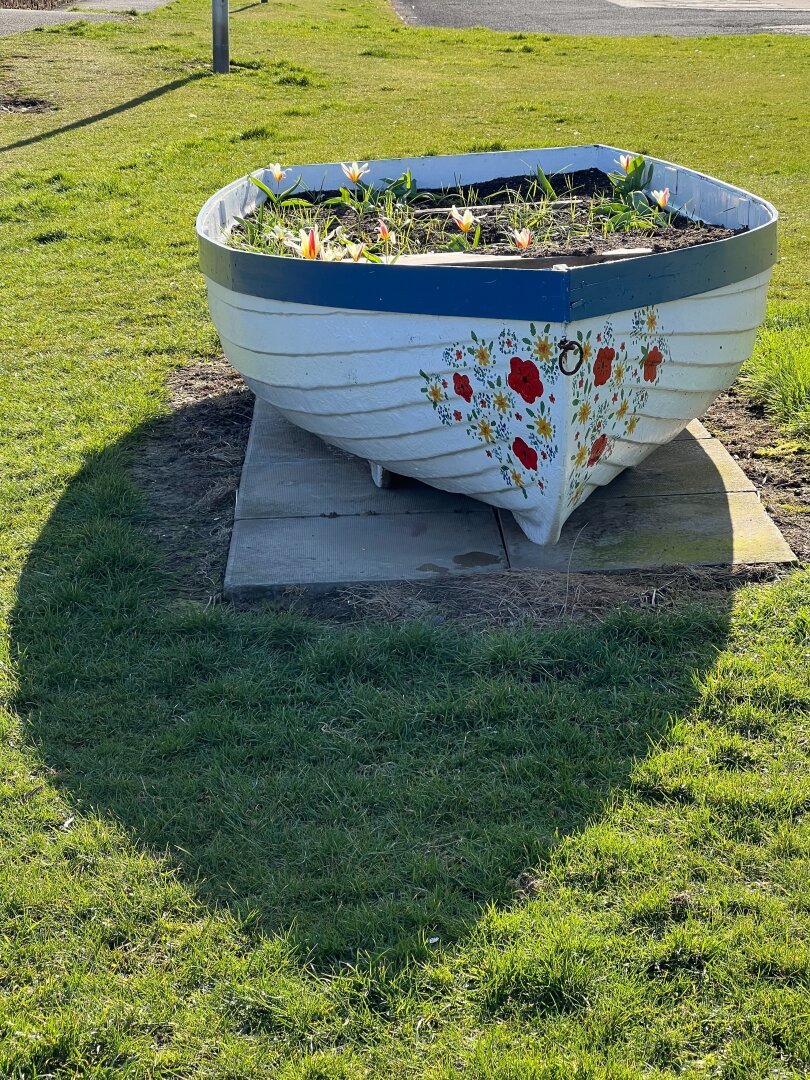 A boat on grass filled with soil and flowers. Flowers painted on the white boat‘s front.