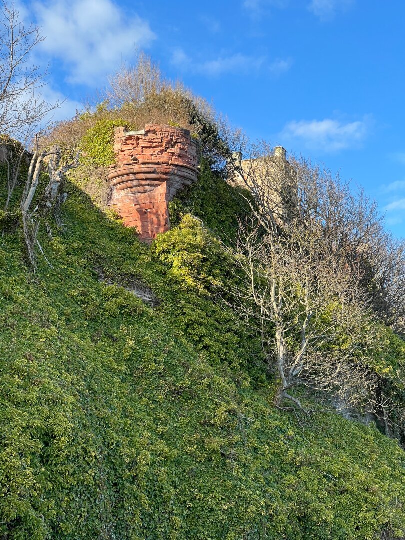 Overgrown cliffs with the remains of a red sandstone castle tower barely visible.