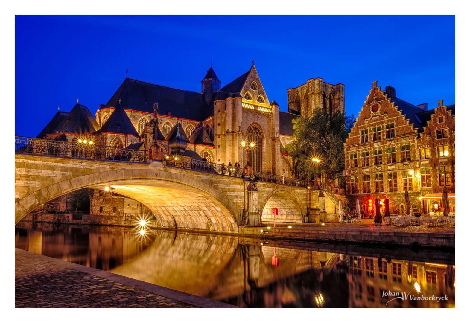 A stone bridge over a river in the foreground with a church in the background under a dark blue night sky. The whole scene is lit up by the yellow city lights.