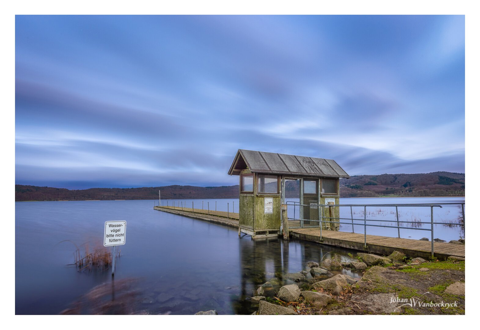 A pier going into a lake under a cloudy sky, with a sign that asks to not feed the birds (in German).