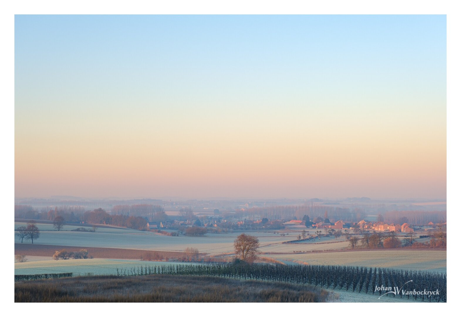 A view over a frozen spring landscape in the morning. There are fields and an orchard in the foreground and some areas with trees and scattered houses in the middle distance and in the background. The view in the distance gets hazy due to a very light fog.