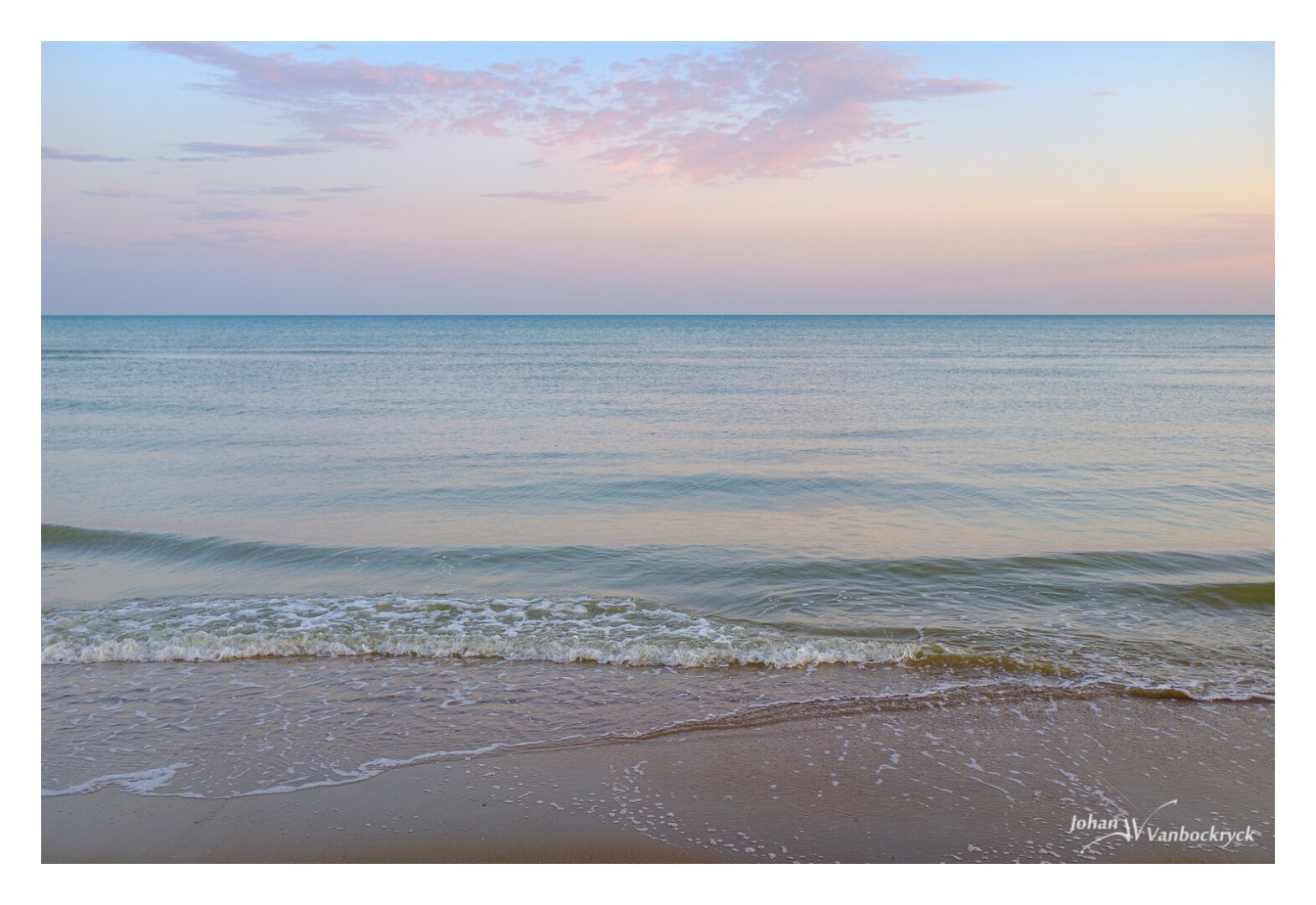 At the edge of the water on the beach, with some low waves coming in under a sky that has a blue, yellow and pink colour from the sunrise.