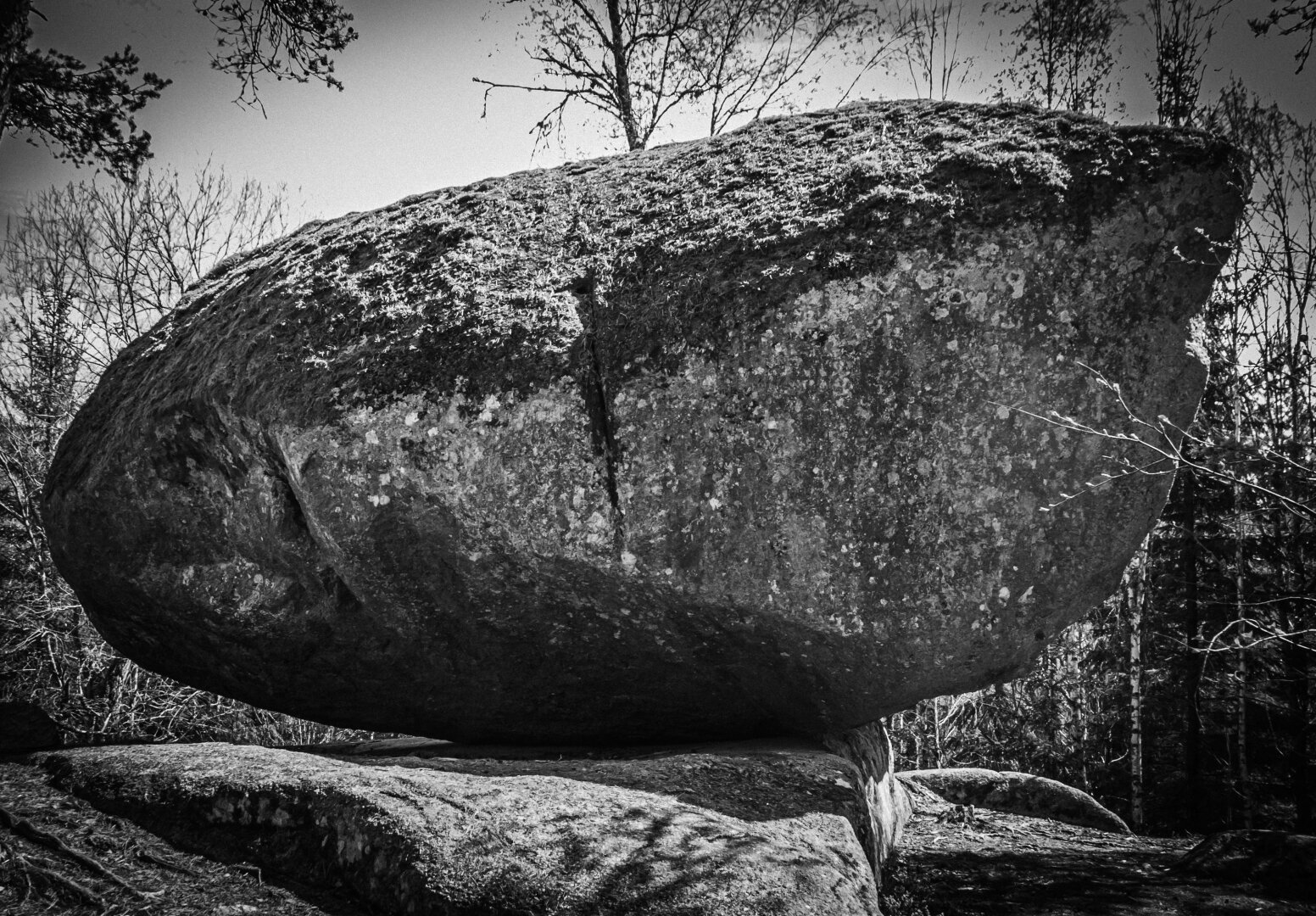 Black-and-white photograph of Runkesten, a massive glacial boulder precariously balanced on a flat rock surface in a leafless forest. The image emphasizes texture, contrast, and the quiet drama of natural equilibrium.