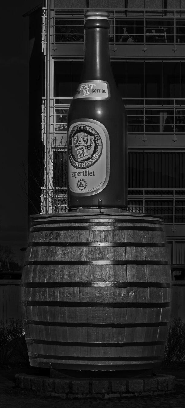 Black-and-white image of a large beer bottle labeled 'FAT 21 GOTT ÖL expertölet' mounted on a wooden barrel, set against an urban backdrop.