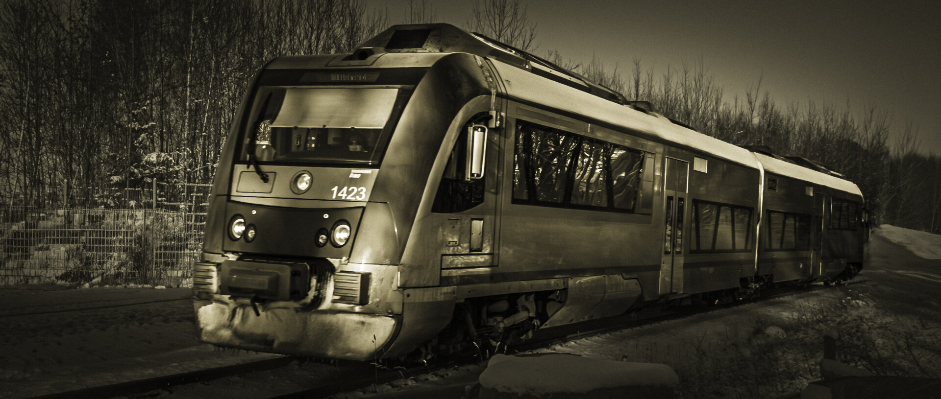 Sepia-toned image of a modern passenger train labeled 1423 moving through a snowy forest landscape. The train contrasts with the leafless trees and snow-covered ground, evoking a sense of quiet transit in winter.
