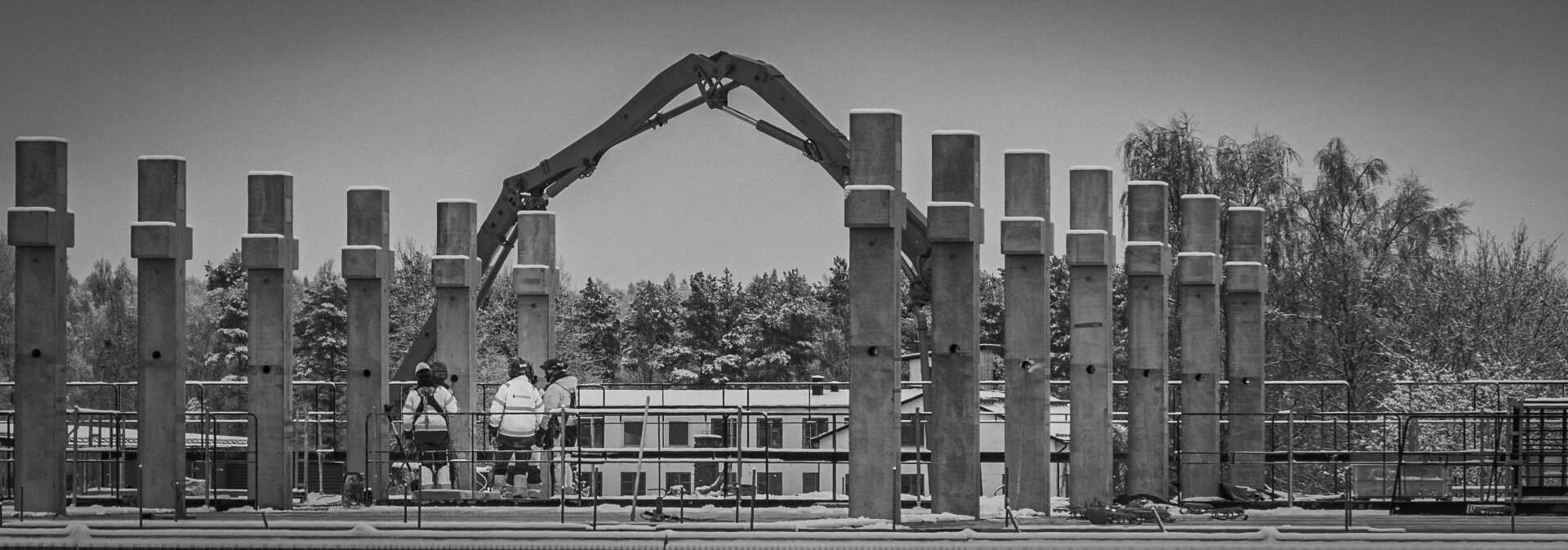 Black-and-white photo of three construction workers standing on a platform between concrete pillars, under a large articulated crane arm. The image has visible grain and vignette, emphasizing structure and mood.