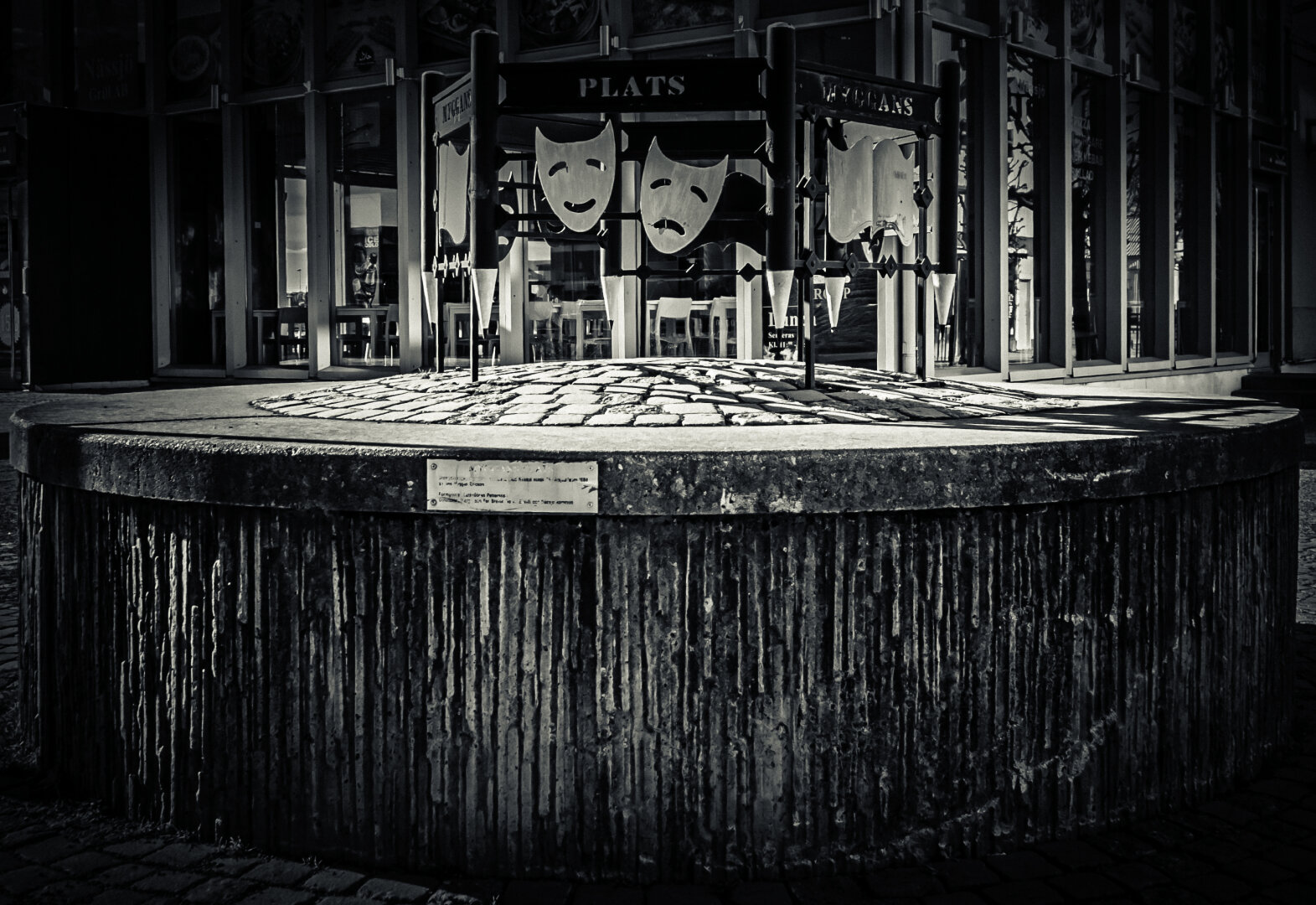 Black and white photo of a circular stone monument with a plaque, in front of a theatrical installation featuring comedy and tragedy masks and the word 'PLATS' above. The scene includes vertical metal poles and a glass-fronted building in the background.