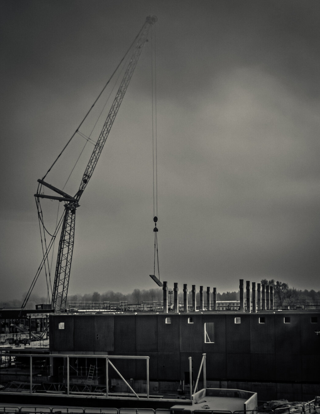 Black-and-white photograph of an industrial construction site. A tall crane stands against a cloudy, foggy sky, lifting a long metal beam. The building below has multiple vertical exhaust pipes and a stark, minimal look. The overall mood is quiet, moody, and atmospheric.