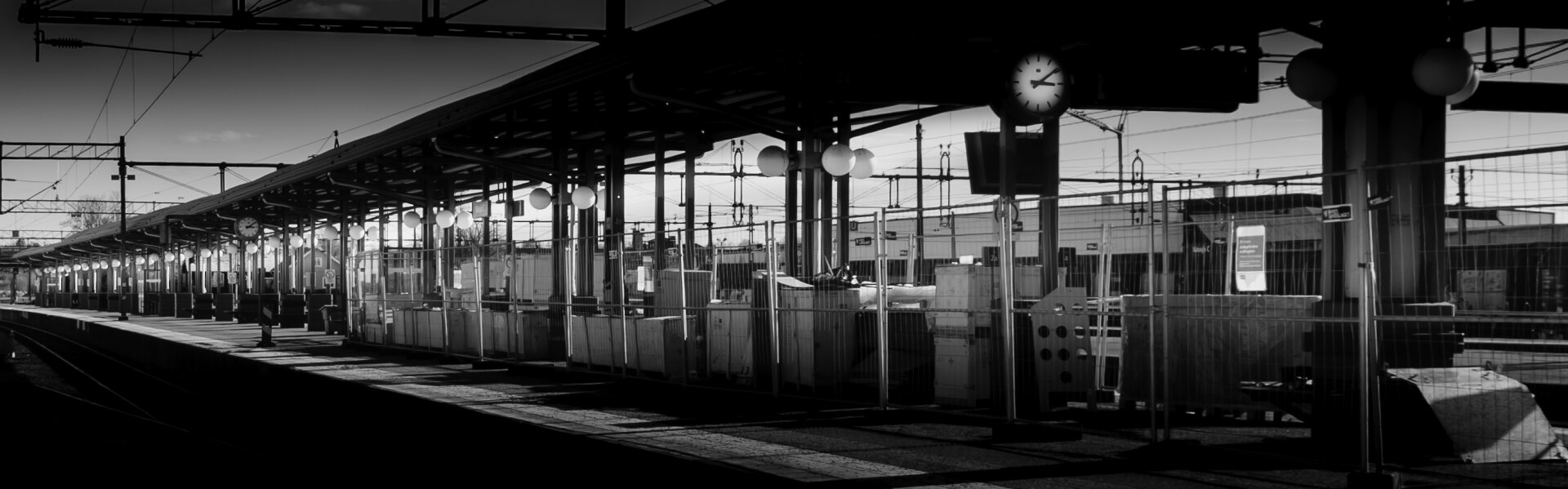 Black-and-white image of a train station platform with vertical beams, overhead wires, and a row of spherical lamps lit along the ceiling. The platform is fenced off, suggesting construction or restricted access. A clock is visible near the center. No people or trains are present.