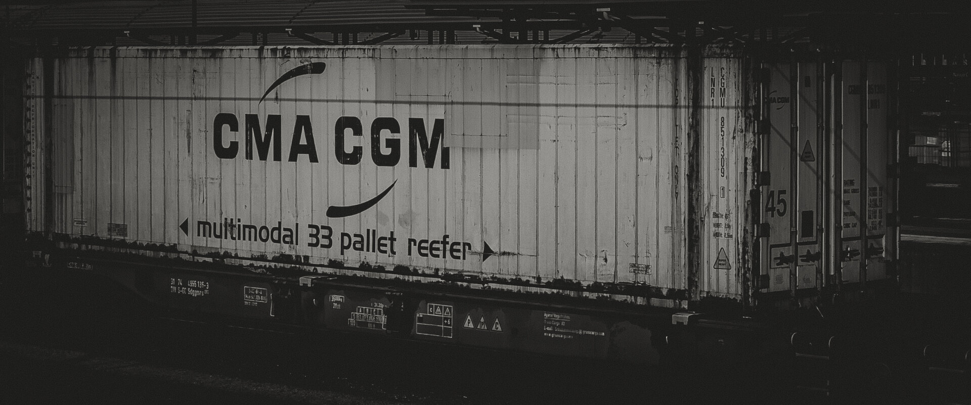 Digitally edited black and white image of a freight train container labeled “CMA CGM multimodal 33 pallet reefer,” mounted on a railcar. The container shows wear and grime. The image mimics Ilford HP5 film with high contrast, visible grain, and a documentary tone.