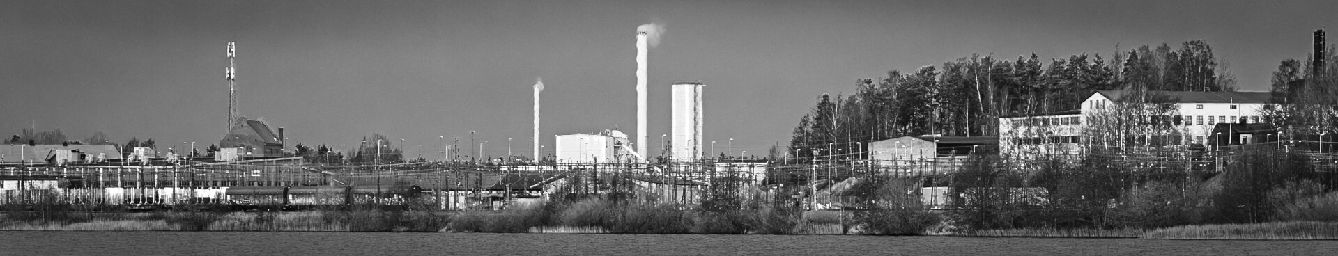 Black-and-white panoramic photo of a district heating power plant near water, with smokestacks releasing smoke, industrial buildings, pipes, and power lines. A forested area is visible in the background under an overcast sky.