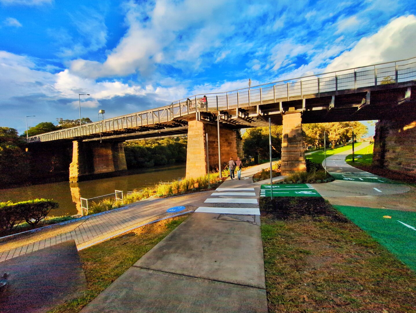 An iron bridge with lattice railings, sitting on wide sandstone pillars. Underneath is a river, a footpath, and a green bike path.