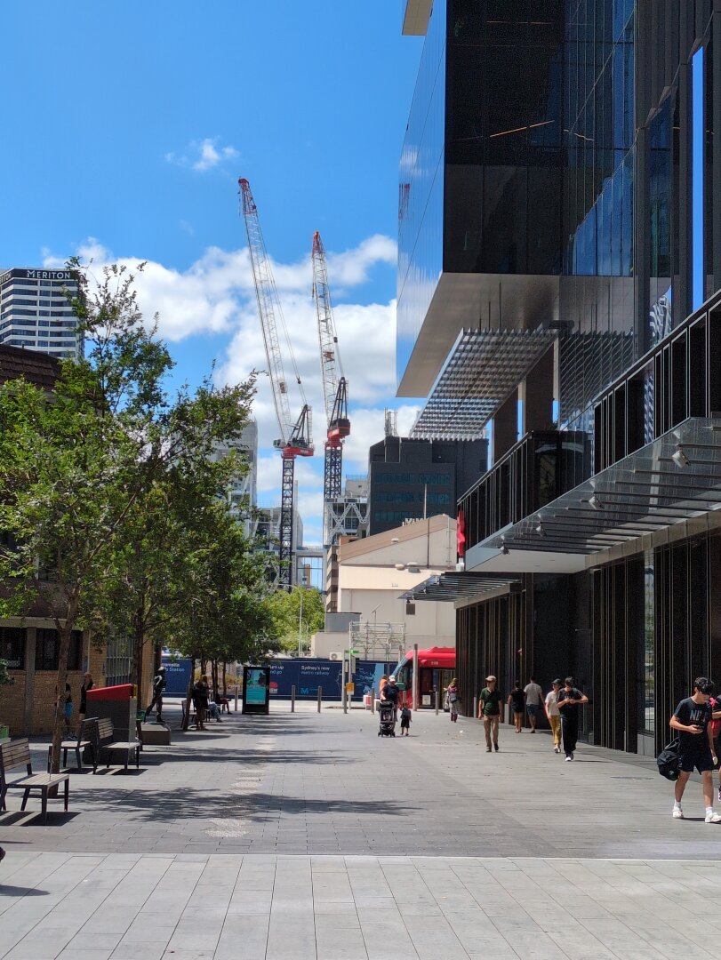 Two cranes on the horizon at the end of a pedestrian alleyway, against a bright blue sky. A tall glass office building stands on the right, and some trees on the left.