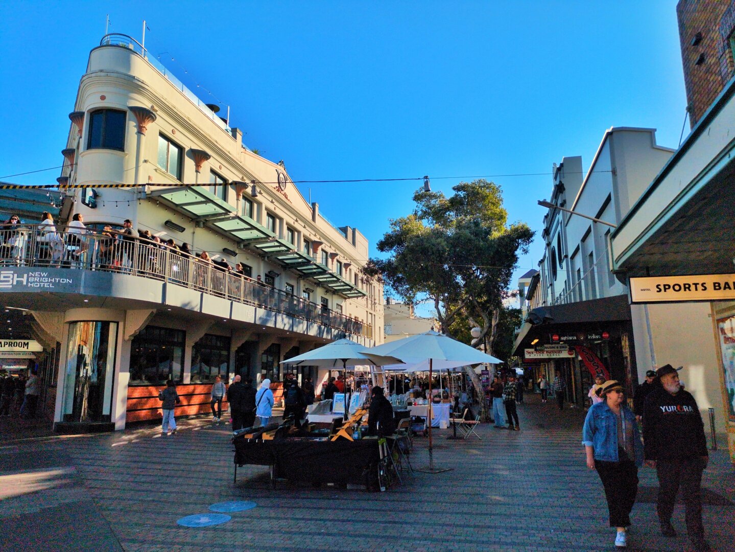 People walk past market stalls along Sydney Rd. The New Brighton Hotel is on the left.