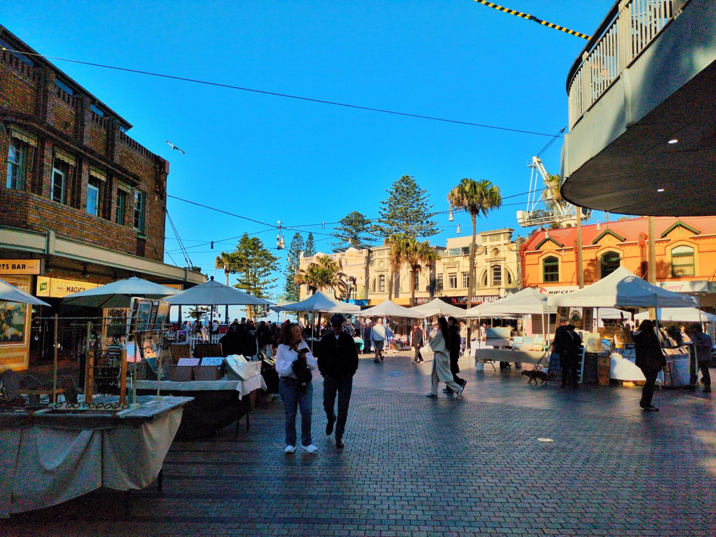 A pedestrianised street with people strolling. It is filled with market stores and shade from the old buildings. There are palm trees and the sky is blue.