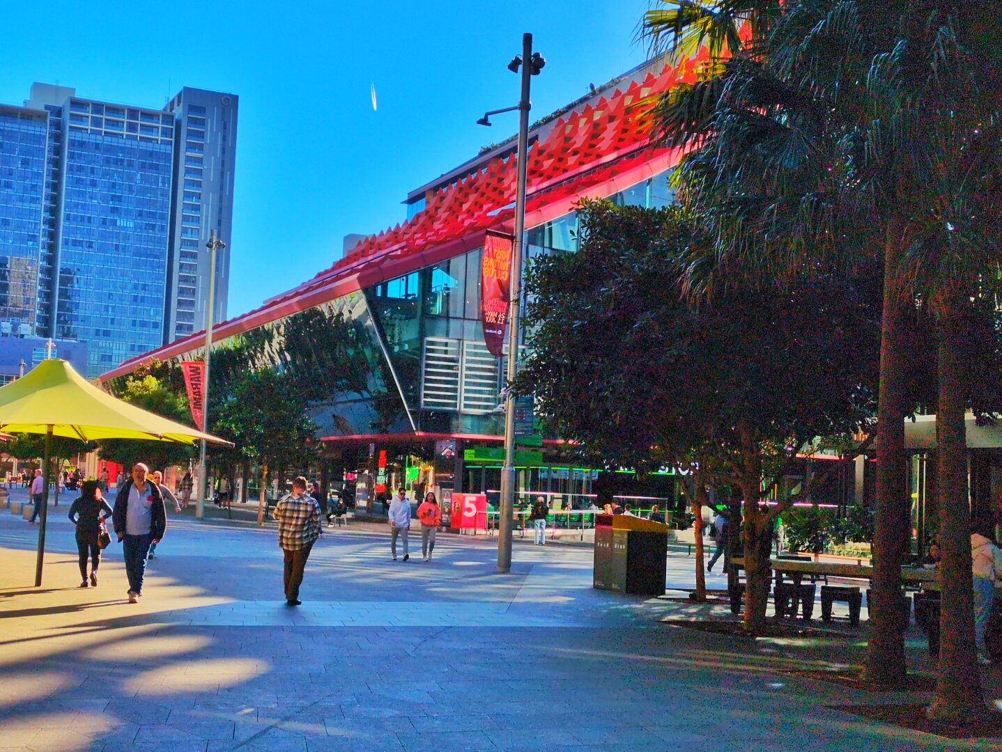The corner of the building at ground level, as viewed from Parramatta Square. People are walking through the square, there's a yellow umbrella and trees.