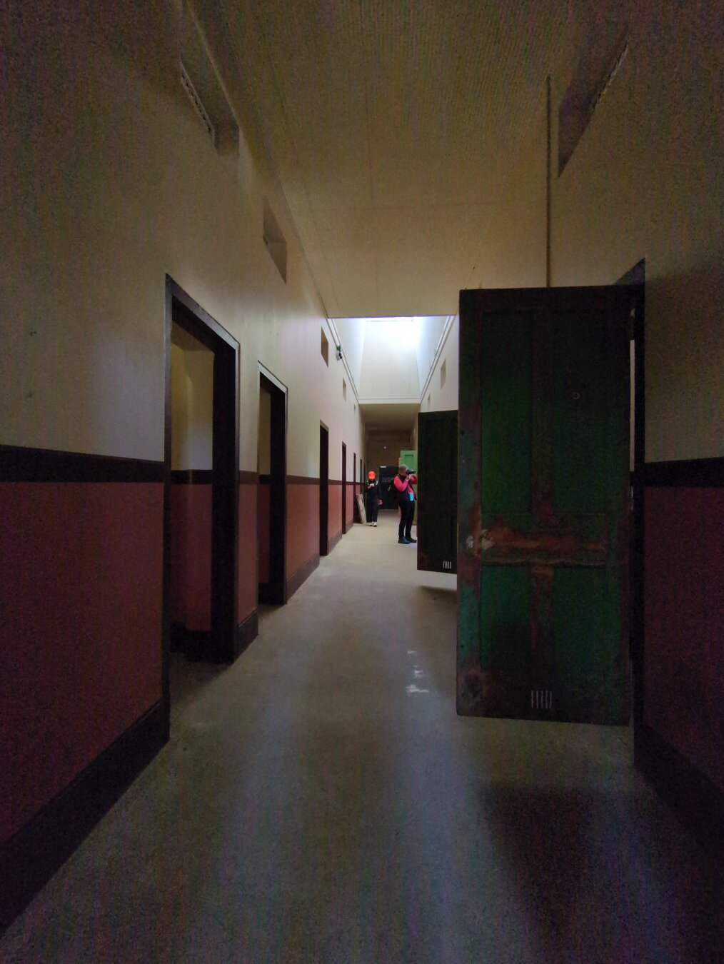 Inside the main corridor, which has the same cream and maroon colour scheme as the previous room. On the right is a row of heavy green steel doors. On the left is a row of rooms with no doors.