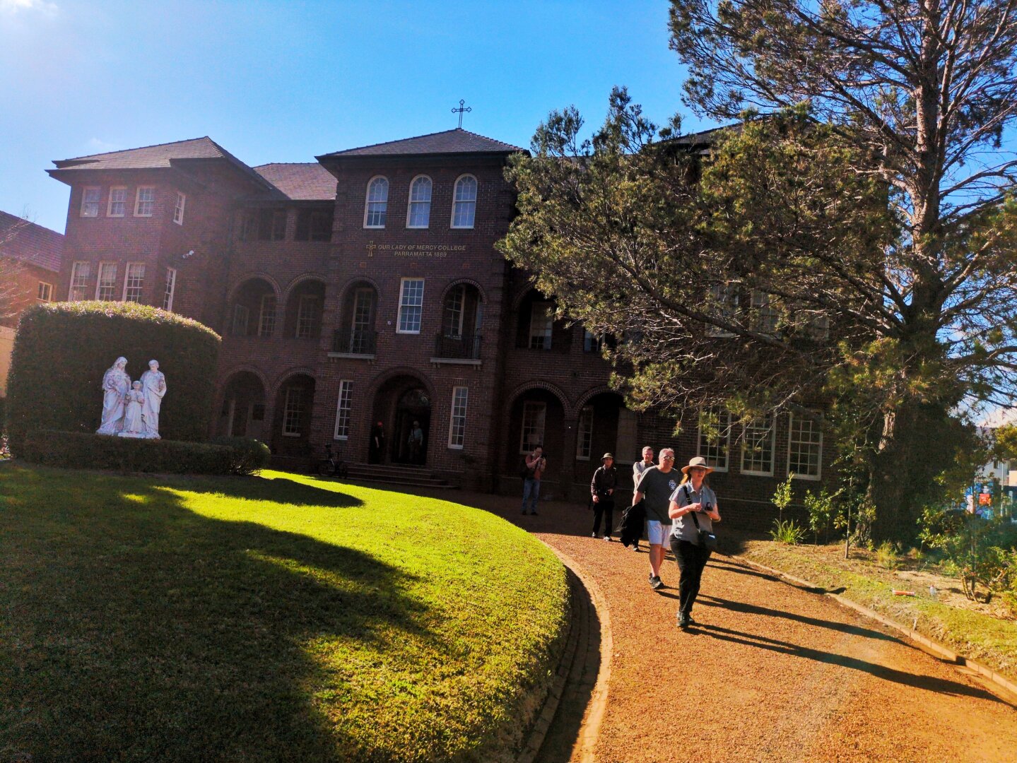 View of the building from the street. There's a path on the right and a lawn with a hedge on the left.