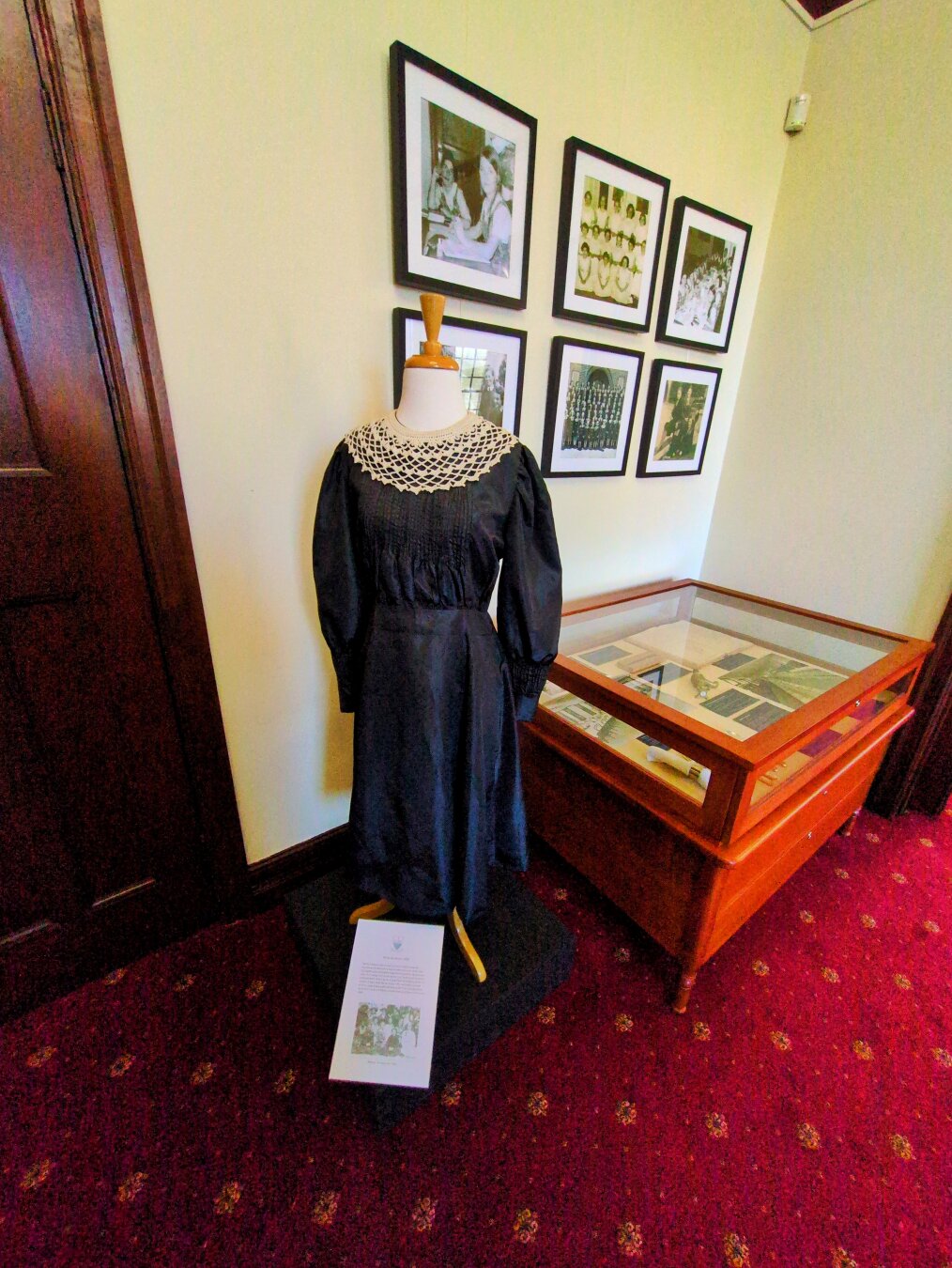 A display near the front office with a collection of photographs of the school in picture frames, a dark dress on a mannequin, and a display case with various records.