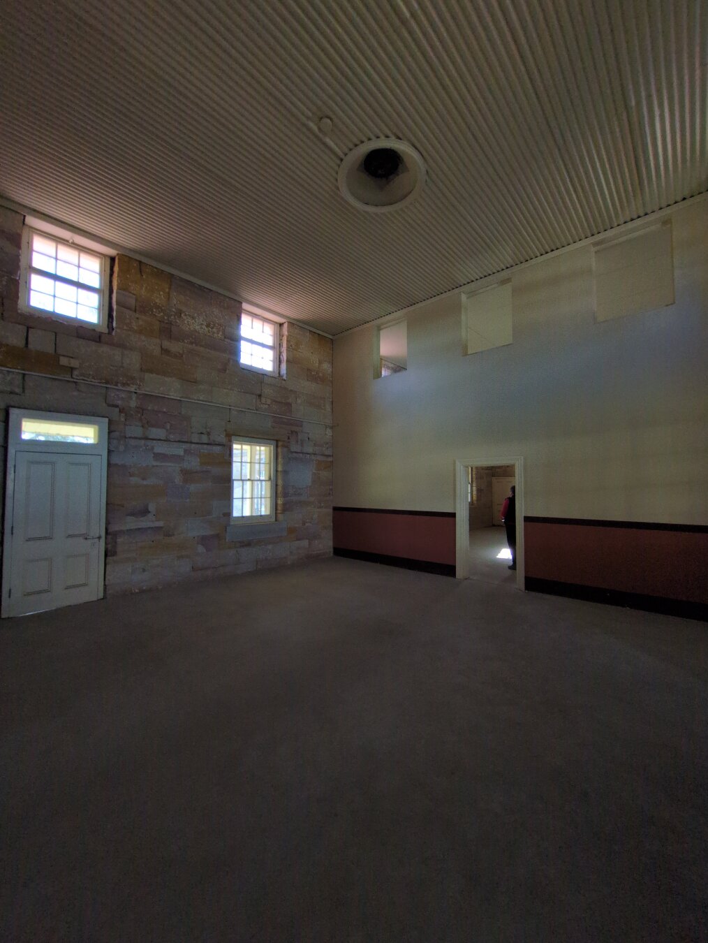 A large entry hall two storeys tall. On the left is a sandstone wall with three windows and a door. On the right in the distance is a cream wall, with the bottom part painted maroon, with a door to the next room. The door looks tiny, but it's actually just a regular door.