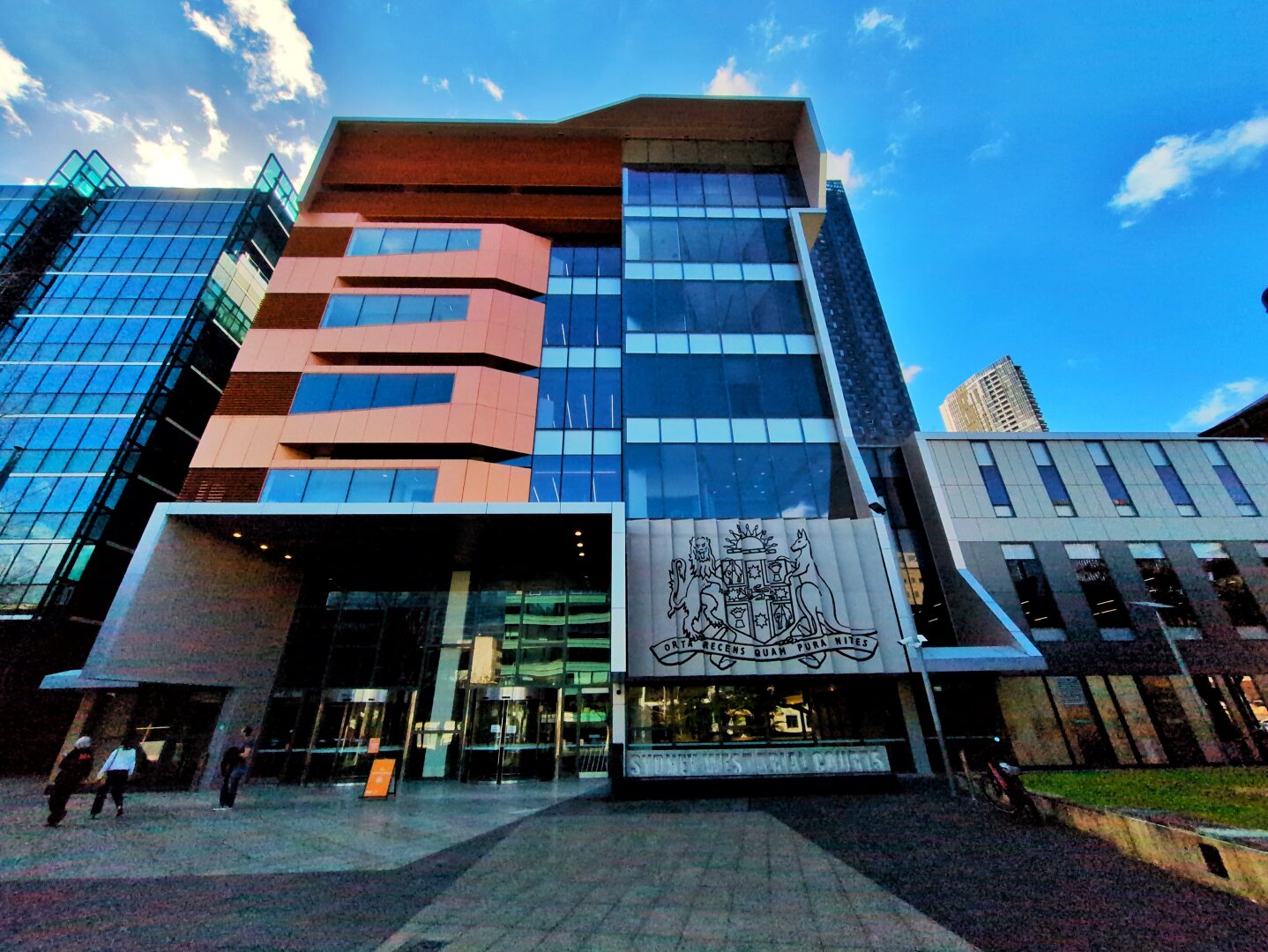 A six-storey modern building. The left of the building is red, while the right is blue, with blue-tinted windows. It has a large metal panel nearly a floor high with the coat of arms of NSW engraved on it.