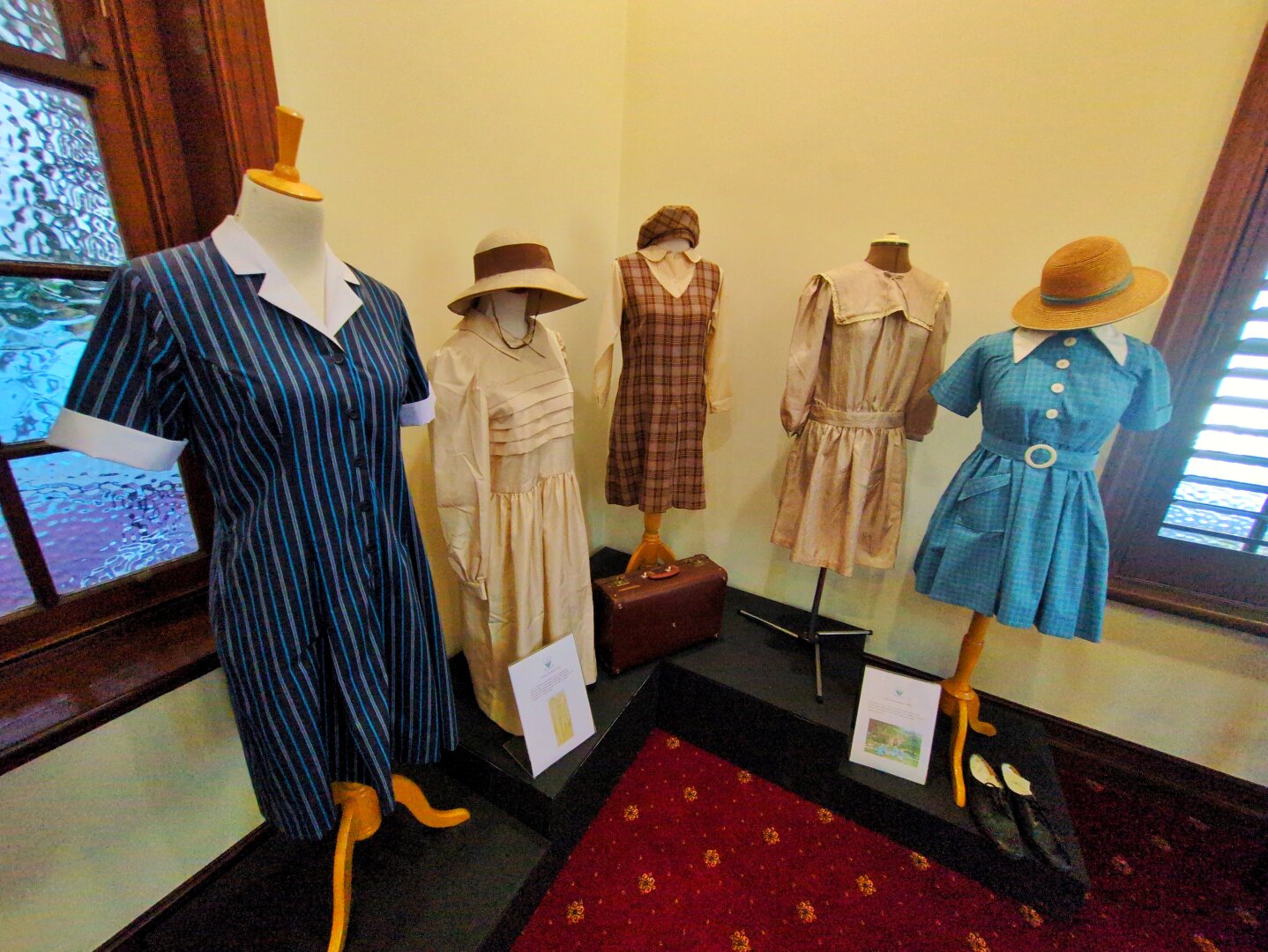 A collection of past uniforms. They range from beige dresses, to brown tartan tunics, to a light blue school dress, and finally a dark blue dress.