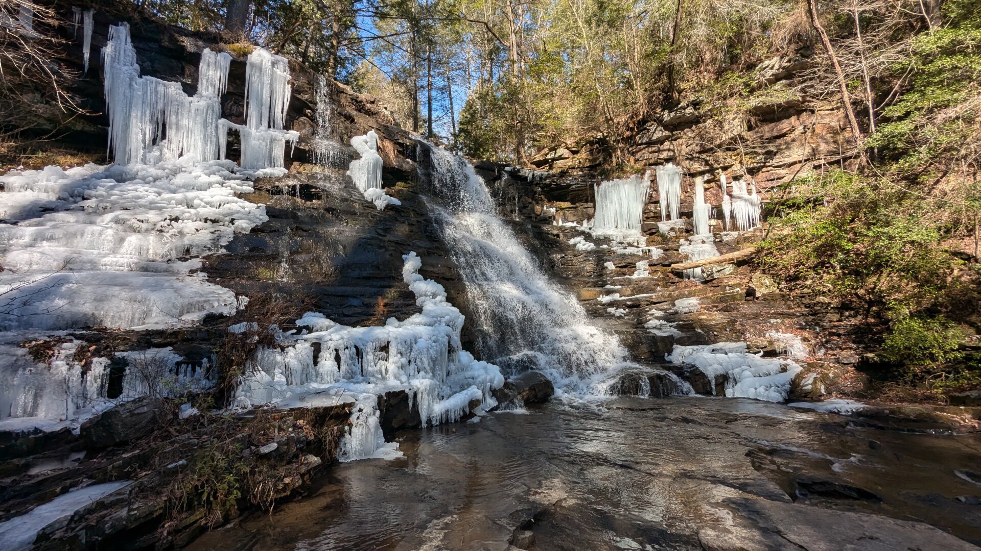 A long bluff with a waterfall flowing in the middle of the frame. on both sides of the waterfall are frozen icicles and i e formations. behind the waterfall is blue sky.