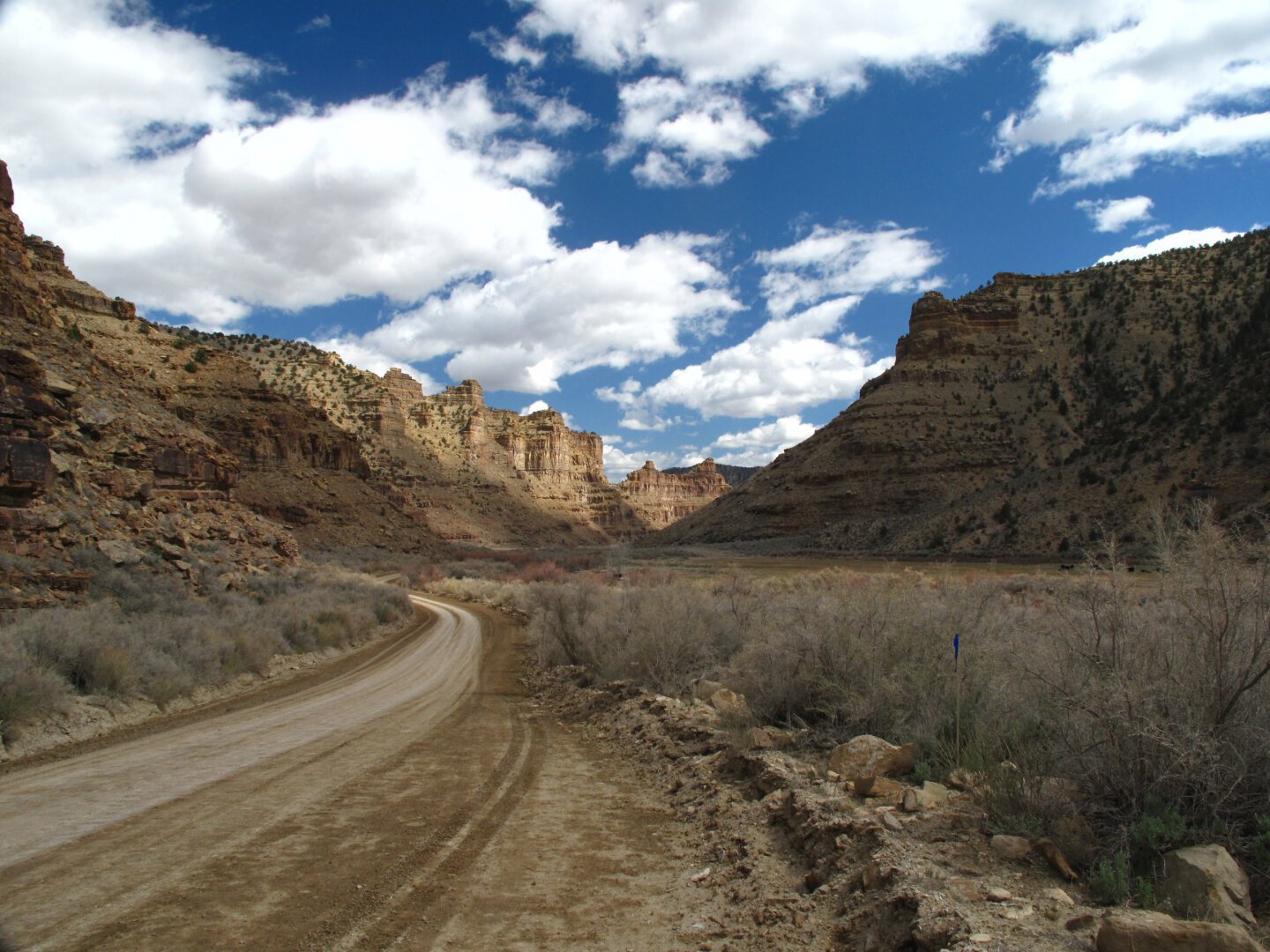 a winding road through a brown desert landscape. On each side are craggy buttes. The sky is bright blue with puffy white clouds.