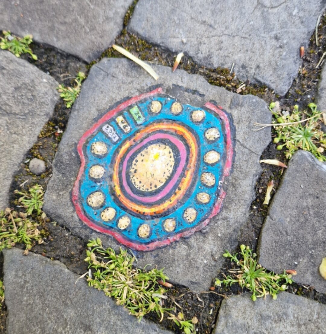 Streetart auf Kopfsteinpglaster: Kreisförmiges Ornament mit blau, weiss und orange, ungefähr so gross wie ein Fünfliber