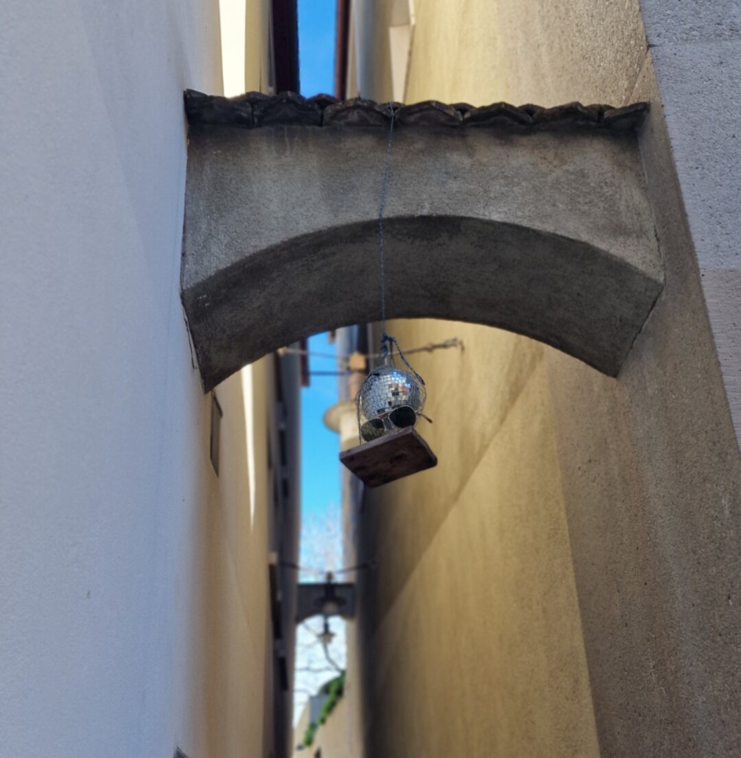 Enge Gasse mit einer Art Brückenverbindung zwischen zwei Häusern. daran hängt eine kleine Discokugel mit Sonnenbrille.
