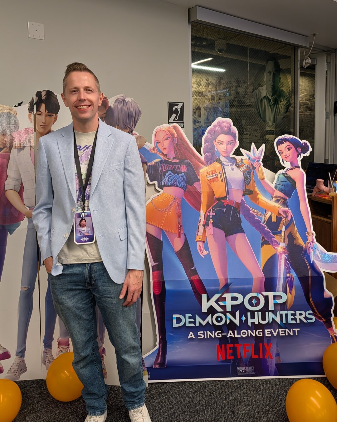 Patrick wearing a Halloween costume of Bobby from K-Pop Demon Hunters, smiles as he stands next to a large promotional cutout for the Netflix movie, K-Pop Demon Hunters: A Sing-Along Event. He is dressed in a light blue blazer over a light gray t-shirt, and blue jeans.

The colorful cardboard cutout features the film's three animated K-pop girl group members, Rumi, Mira, and Zoey (HUNTR/X), wielding swords and standing in front of the male demon boy band (Saja Boys). Black and yellow balloons decorate the floor near the display. Patrick is also wearing a lanyard with a badge that appears to say "VIP."